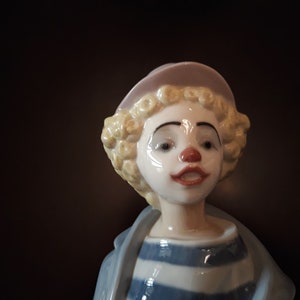 May include: A porcelain figurine of a clown with blonde curly hair wearing a pink hat and a blue and white striped shirt.