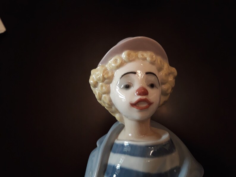 May include: A porcelain figurine of a clown with blonde curly hair wearing a pink hat and a blue and white striped shirt.