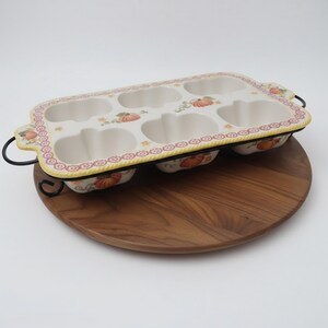 May include: A rectangular ceramic baking pan with six pumpkin-shaped cavities, featuring a cream-colored interior and a decorative border with orange pumpkin motifs. The pan is set in a black metal wire frame with side handles for easy transport.