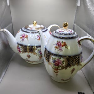 May include: Two white porcelain teapots featuring intricate floral patterns in pink, orange, and purple. Each teapot is accented with deep blue and gold filigree detailing, a curved handle, a spout, and a gold-toned knob on the lid.