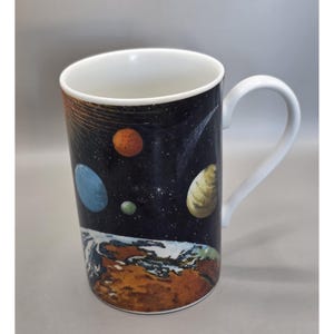 May include: White ceramic mug featuring a colorful illustration of planets, stars, and Earth against a black background. The mug has a white handle and interior, perfect for coffee or tea.