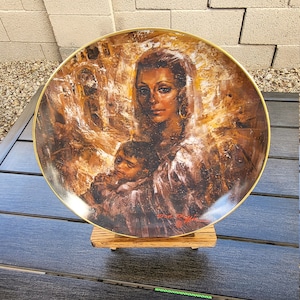 Pode incluir: Um prato de cerâmica decorativo com uma borda dourada apresenta uma pintura de uma mulher e uma criança. A mulher está usando um véu branco e tem cabelo escuro e olhos castanhos. A criança está usando um casaco marrom e tem cabelo escuro. O fundo é uma pintura desfocada e abstrata de uma cena da cidade.