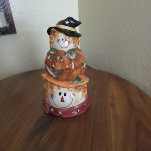 May include: A ceramic figurine featuring a stack of three elements: a top figure wearing a black witch hat, a middle orange jack-o'-lantern with cut-out facial features, and a bottom figure with a smiling face and a maroon scarf base.