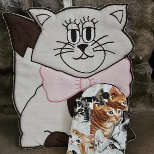 May include: A white cat-shaped fabric wall hanging with a pink bow and a brown ear. The cat has embroidered eyes and whiskers. A smaller fabric pouch with a cat print is hanging from the cat's neck.