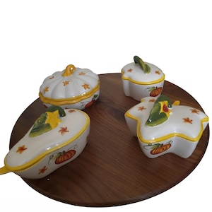 May include: A set of four white ceramic lidded containers with yellow rims and painted pumpkin motifs. The pieces include a round pumpkin-shaped dish, a pear-shaped dish, a star-shaped dish, and a small round dish, all featuring green stems.