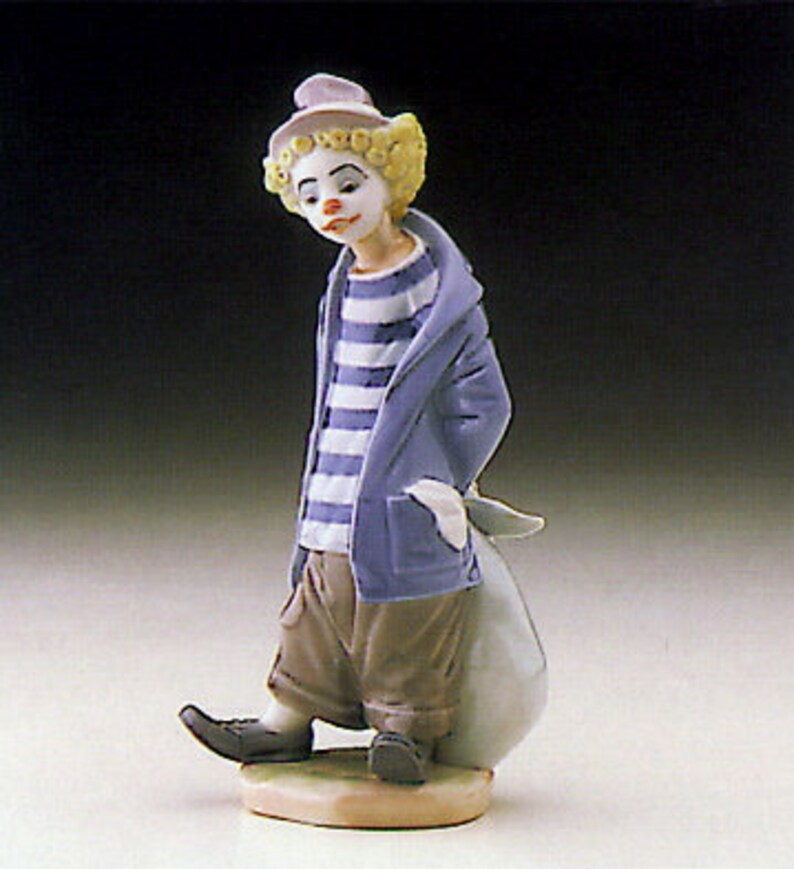 May include: A porcelain figurine of a clown with a blue and white striped shirt, a blue jacket, and a white hat. The clown is holding a white bag and is standing on a small base.