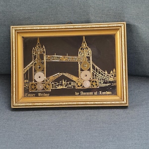 Op de afbeelding: Een ingelijst kunstwerk met een goudkleurige afbeelding van de Tower Bridge tegen een zwarte achtergrond. Het brugontwerp bevat tandwielen en andere mechanische elementen. De tekst "Tower Bridge by Ammon of London" is zichtbaar.