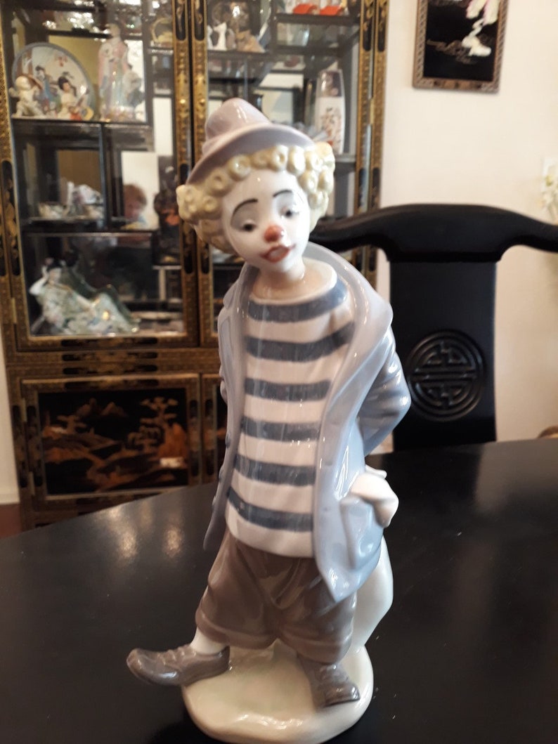 May include: A porcelain figurine of a clown with a blue and white striped shirt, a gray jacket, and brown pants. The clown is standing on a white base.
