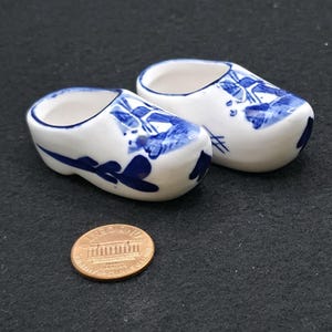 May include: Two miniature ceramic clogs, painted white with blue floral designs. The clogs are next to a penny, providing a size reference. The clogs are a traditional Dutch souvenir.