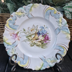 May include: A white porcelain plate with a scalloped edge, featuring a hand-painted floral design. The flowers are in shades of yellow, pink, purple, and blue, with gold trim accents. The plate is displayed on a decorative metal stand.
