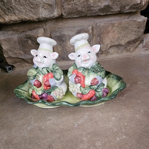 May include: A ceramic salt and pepper set featuring two smiling pig figurines dressed as chefs. Each pig holds vegetables and wears a chef's hat. The set sits on a green, leaf-shaped dish. The pigs are white with pink snouts and ears.