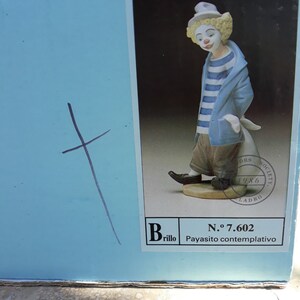May include: A porcelain figurine of a clown with a blue and white striped shirt and a white hat. The figurine is standing on a base with the text "N.&ordm; 7.602 Payasito contemplativo" and "Brillo".