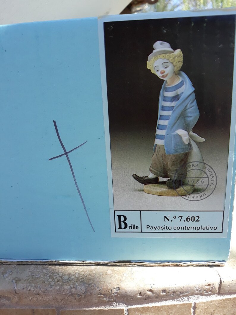 May include: A porcelain figurine of a clown with a blue and white striped shirt and a white hat. The figurine is standing on a base with the text "N.&ordm; 7.602 Payasito contemplativo" and "Brillo".