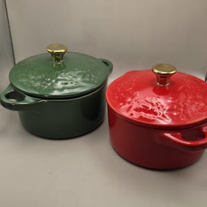 Może przedstawiać: Dwa małe, okrągłe ceramiczne garnki z pokrywkami. Jeden jest ciemnozielony, drugi czerwony. Oba mają złote gałki na pokrywkach i małe uchwyty po bokach. Pokrywki mają teksturowany, wytłoczony wzór.