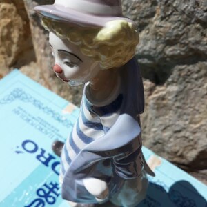 May include: A porcelain figurine of a clown with a blue and white striped shirt, a blue jacket, and a purple hat. The clown is looking down and has a sad expression.
