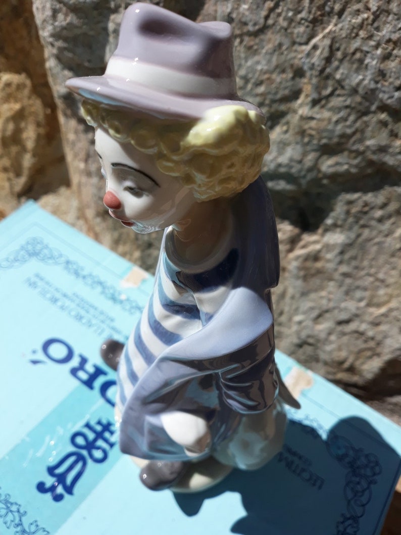 May include: A porcelain figurine of a clown with a blue and white striped shirt, a blue jacket, and a purple hat. The clown is looking down and has a sad expression.