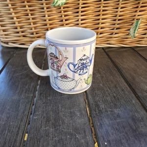 Könnte beinhalten: Weißer Keramikbecher mit verschiedenen Teekannen-Designs in Rosa, Blau und Grün. Der Becher hat einen weißen Henkel und einen hellblau gestreiften Hintergrund mit floralen Akzenten. Eine charmante Ergänzung für jede Küche.