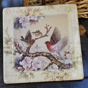 Könnte beinhalten: Eine quadratische Keramikfliese mit einem Blumendesign und drei Vögeln. Die Vögel sitzen auf einem Ast mit rosa Blüten. Die Fliese trägt das Wort "Harmonie" viermal.
