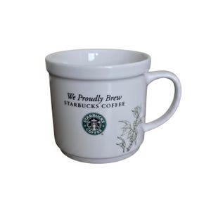May include: A white ceramic mug featuring the text We Proudly Brew STARBUCKS COFFEE in black lettering. Below the text is the circular green and white Starbucks Coffee logo. A grey botanical illustration of coffee branches is on the right side.