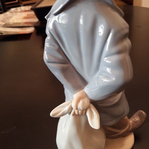 May include: A porcelain figurine of a person wearing a blue coat and holding a white bag. The figurine is standing on a white base.