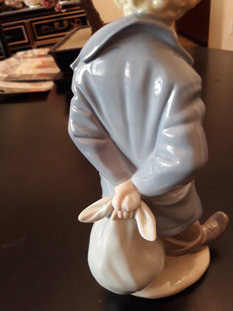 May include: A porcelain figurine of a person wearing a blue coat and holding a white bag. The figurine is standing on a white base.