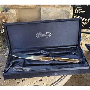 Könnte beinhalten: Ein silberfarbenes Tranchiermesser mit goldfarbenem Griff liegt in einer offenen, marineblauen, samtgefütterten Schatulle. Die Innenseite der Schatulle ist mit Satin ausgekleidet. Auf dem Deckel steht "Things Remembered".