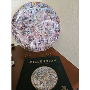 May include: A decorative plate with a spiral design composed of historical images, displayed on a gold-colored stand. The plate is resting on a wooden surface next to a book titled "The Millennium Time Tapestry".