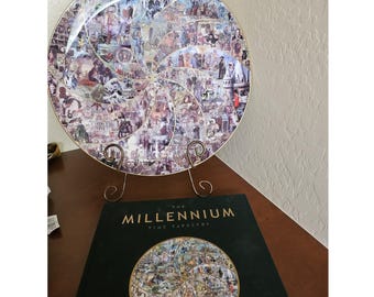 May include: A decorative plate with a spiral design composed of historical images, displayed on a gold-colored stand. The plate is resting on a wooden surface next to a book titled "The Millennium Time Tapestry".