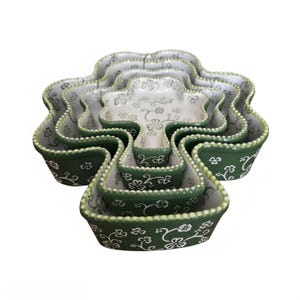 May include: A set of four nested clover-shaped bowls in varying sizes. Each bowl features a deep green exterior and a white interior, both decorated with a white and green floral vine pattern. The rims are finished with a raised beaded texture.