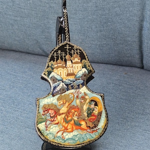 May include: A black, decorative, violin-shaped figurine with a painted winter scene. The figurine features a church, horses pulling a sleigh, and intricate gold detailing. The base is round and black.