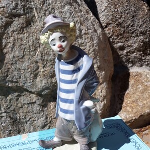 May include: A porcelain figurine of a clown with a blue and white striped shirt, a blue jacket, and a white hat. The clown is holding a white bag and is standing on a white base. The figurine is on a blue box with the word "DRO" printed on it.