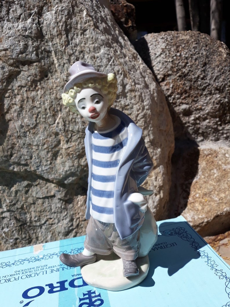 May include: A porcelain figurine of a clown with a blue and white striped shirt, a blue jacket, and a white hat. The clown is holding a white bag and is standing on a white base. The figurine is on a blue box with the word "DRO" printed on it.