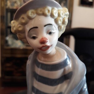 May include: A porcelain figurine of a clown with a white face, red nose, and blonde curly hair. The clown is wearing a blue and white striped shirt and a lavender hat.