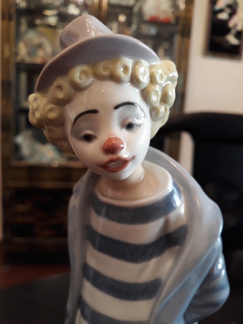 May include: A porcelain figurine of a clown with a white face, red nose, and blonde curly hair. The clown is wearing a blue and white striped shirt and a lavender hat.