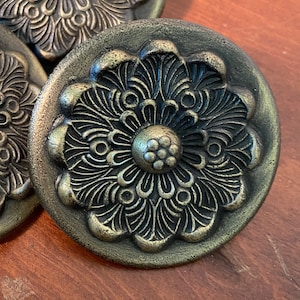May include: Three circular metal buttons featuring an antique brass finish and a raised, intricate floral pattern. Each button displays a central cluster of small dots surrounded by radiating, detailed petals that extend to the outer edge of the disc.