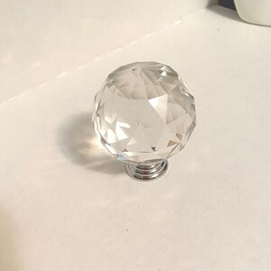 May include: A clear, faceted crystal cabinet knob with a chrome base. The knob is spherical and reflects light, creating a sparkling effect. This decorative hardware is suitable for drawers and cabinets.