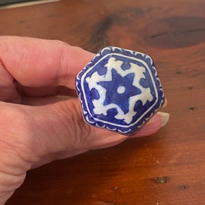 May include: A blue and white ceramic knob with a hexagonal shape and a star design. The knob has a white background with a blue star in the center and a blue border.