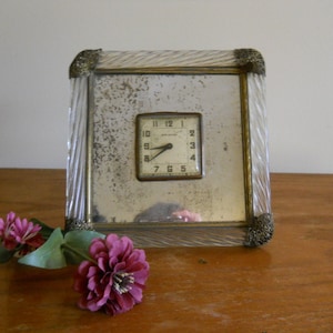 May include: A vintage square clock with a mirrored face and a small clock face. The clock is framed in clear, ridged glass with ornate corner details. A pink flower with green leaves rests on the wooden surface in front of the clock.