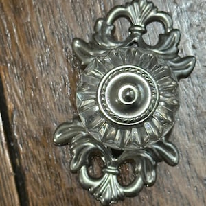 May include: A clear glass cabinet knob with a decorative silver-toned backplate. The knob has a circular, faceted design, and the backplate features an ornate, floral-inspired pattern. The hardware is set against a wooden surface.