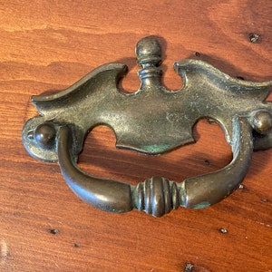 May include: A vintage-style, ornate brass drawer pull with a decorative, stylized design. The pull has a curved handle and a decorative top.