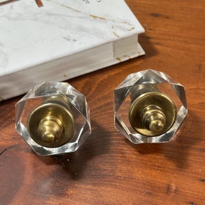 May include: Two clear, faceted crystal cabinet knobs with brass hardware. The knobs are hexagonal and sit on a wooden surface next to a book with a marble-like cover. These decorative knobs are ideal for furniture upgrades.