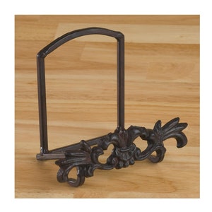 May include: A dark brown metal plate stand with a fleur-de-lis design. The stand has a curved top and a decorative base. The plate holder is made of cast iron and is designed to hold a plate or other decorative item.