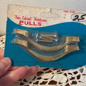 May include: Vintage cabinet pulls, packaged on a blue and white card. The card reads "Fine Cabinet Hardware PULLS." The pulls are gold-toned, with a curved design. The package includes screws for installation. The card is marked "Made in Japan."
