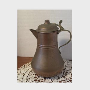 May include: A copper-colored pitcher with a lid and a curved handle. The pitcher has a spout for pouring and a decorative handle. The pitcher is sitting on a white lace doily.