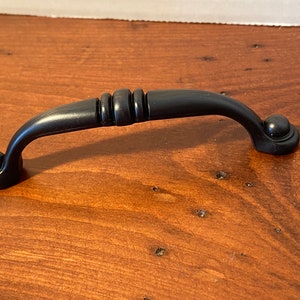 May include: Black cabinet pull with a rounded design and a decorative detail in the center. The pull has a smooth, matte finish.