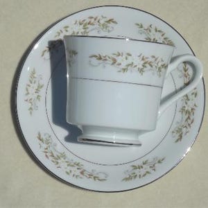 May include: A white teacup and saucer with a floral pattern in shades of brown and green. The saucer has a scalloped edge.