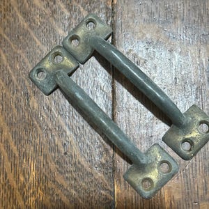 May include: Two antique metal drawer pulls with a brass finish on the mounting plates and a dark gray handle. The pulls are rectangular with three holes for screws and a curved handle. The background is a wooden surface.