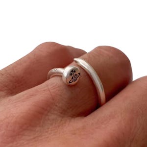 May include: A silver-colored ring with a nail-head design. The ring features a rounded head with a black and white emblem and a curved band that wraps around the finger. The ring is worn on a finger.
