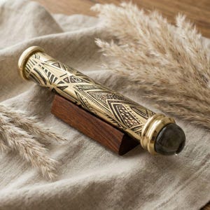 May include: A gold-colored kaleidoscope with black geometric patterns rests on a small wooden stand. The kaleidoscope has a gemstone at one end and is displayed on a neutral-toned linen surface.