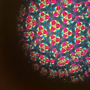 May include: Kaleidoscope image with a repeating pattern of colorful, geometric shapes. The design features vibrant pink, yellow, and blue hues against a dark background. The pattern creates a symmetrical, abstract design, reminiscent of a flower or star.